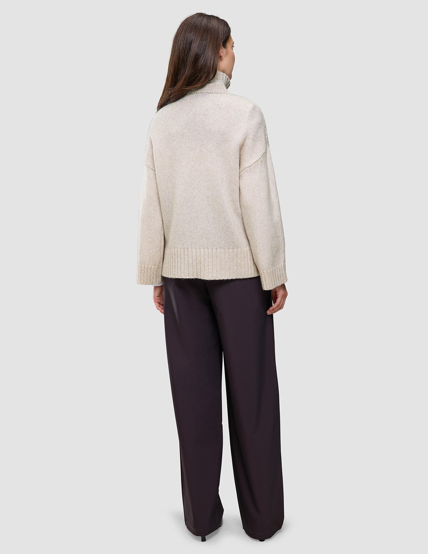 Eine Person, von hinten gesehen, die einen cremefarbenen, dicken, weichen Damen-Rollkragenpullover aus Wolle in der Farbe Sugar Melange sowie eine dunkle Hose mit weitem Bein trägt.
